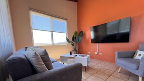 Ein Wohnzimmer mit zwei grauen Sofas, einem weißen Couchtisch, einem Flachbildfernseher an einer orangefarbenen Wand und einem Fenster mit Jalousien.