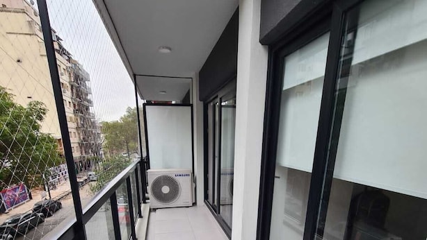 En smal balkon med en Samsung-aircondition, en seng og udsigt til gaden.
