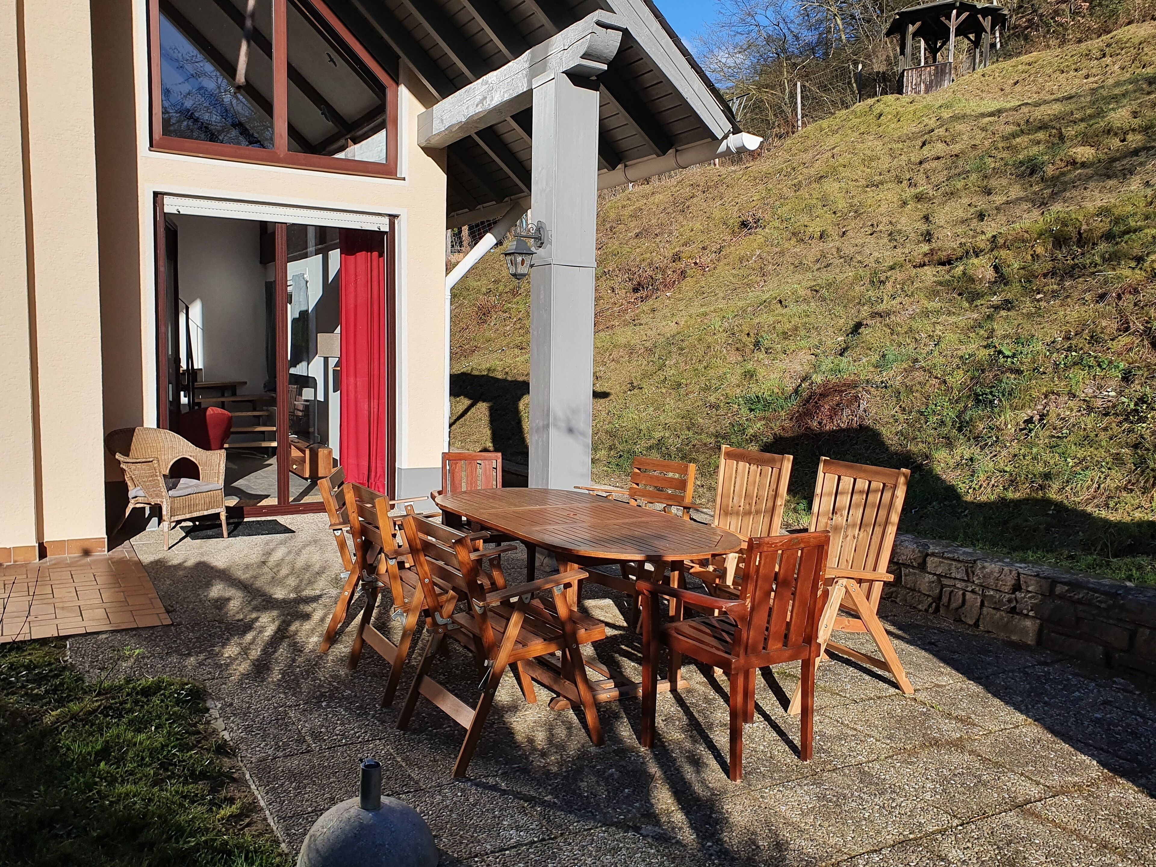An outdoor patio with a wooden table and chairs, a red curtain, and a view of a hillside.