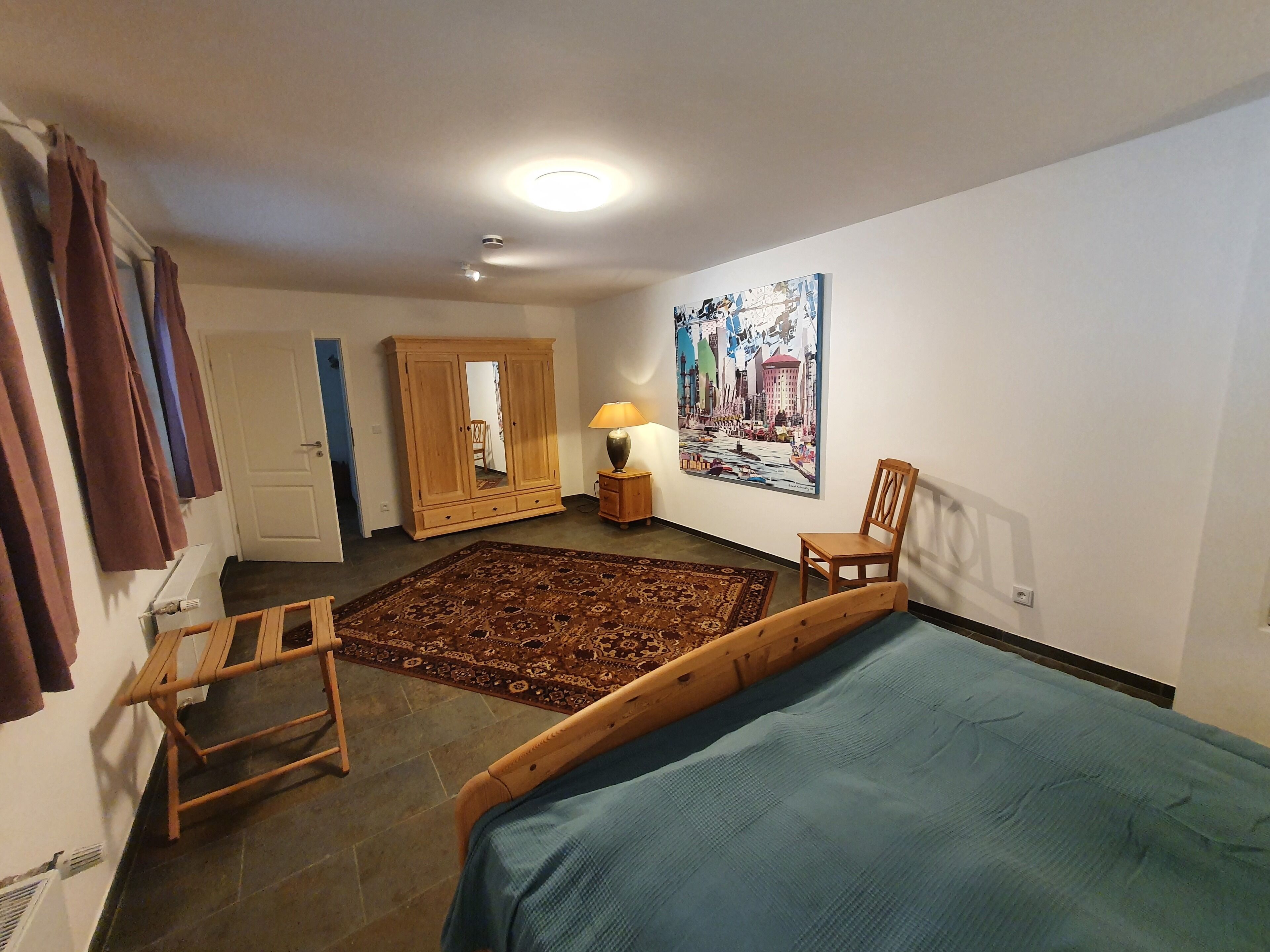 Een slaapkamer met een bed, een houten stoel, een kledingkast, een vloerkleed, een lamp en een stadslandschap schilderij.