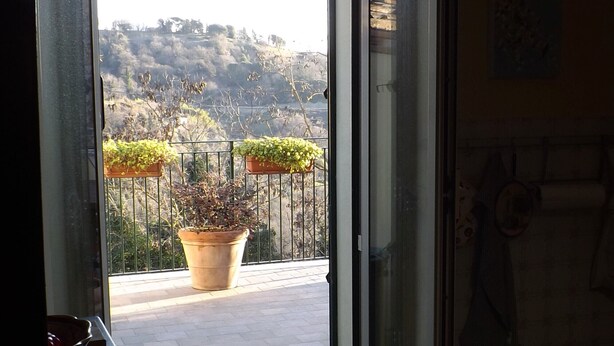 Une porte ouverte donnant sur un balcon agrémenté de plantes en pot et offrant une vue sur un paysage vallonné.