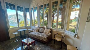 Ein Zimmer mit einer Couch, Sesseln, einem kleinen Tisch und großen Fenstern mit Blick auf Palmen und ein fernes Gebirge.