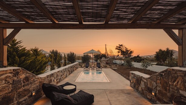 Zona de piscina con muros de piedra, sillas de descanso y vista a un edificio lejano al atardecer.