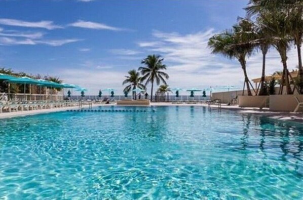 A clear blue pool with palm trees and umbrellas, surrounded by a fence. - Beachfront Resort-style Living! (Hollywood)