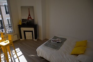 A bedroom with a bed, a small table, a chair, a fireplace with a painting of the Eiffel Tower, and a balcony with a railing.