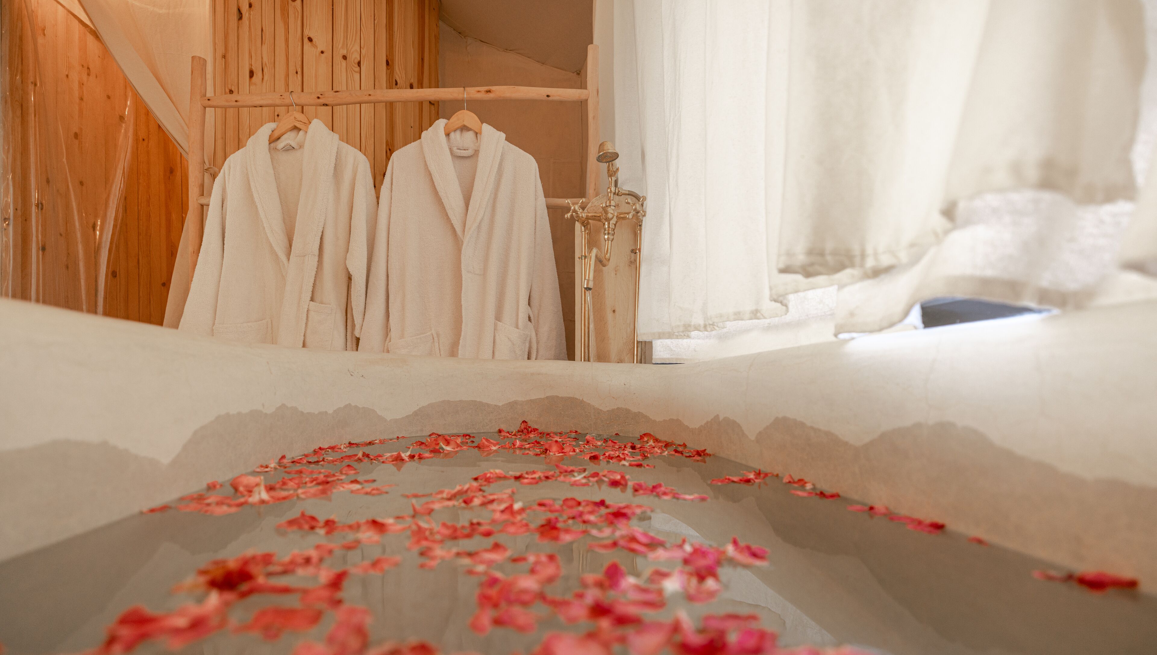 Un baño con paredes de madera, una bañera con pétalos de rosa y dos albornoces blancos colgados en un perchero.