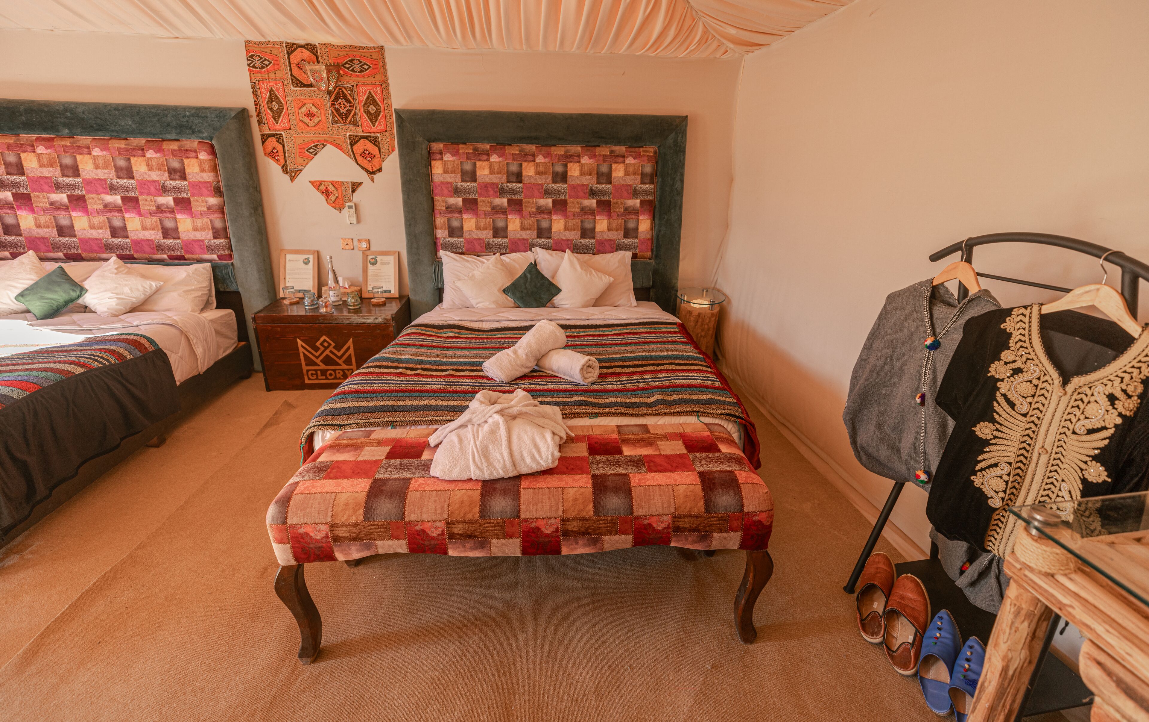 Ein Hotelzimmer mit zwei Betten, einer Holzkistenkommode, einem Kleidungsstück zum Aufhängen und einem Paar Hausschuhen.