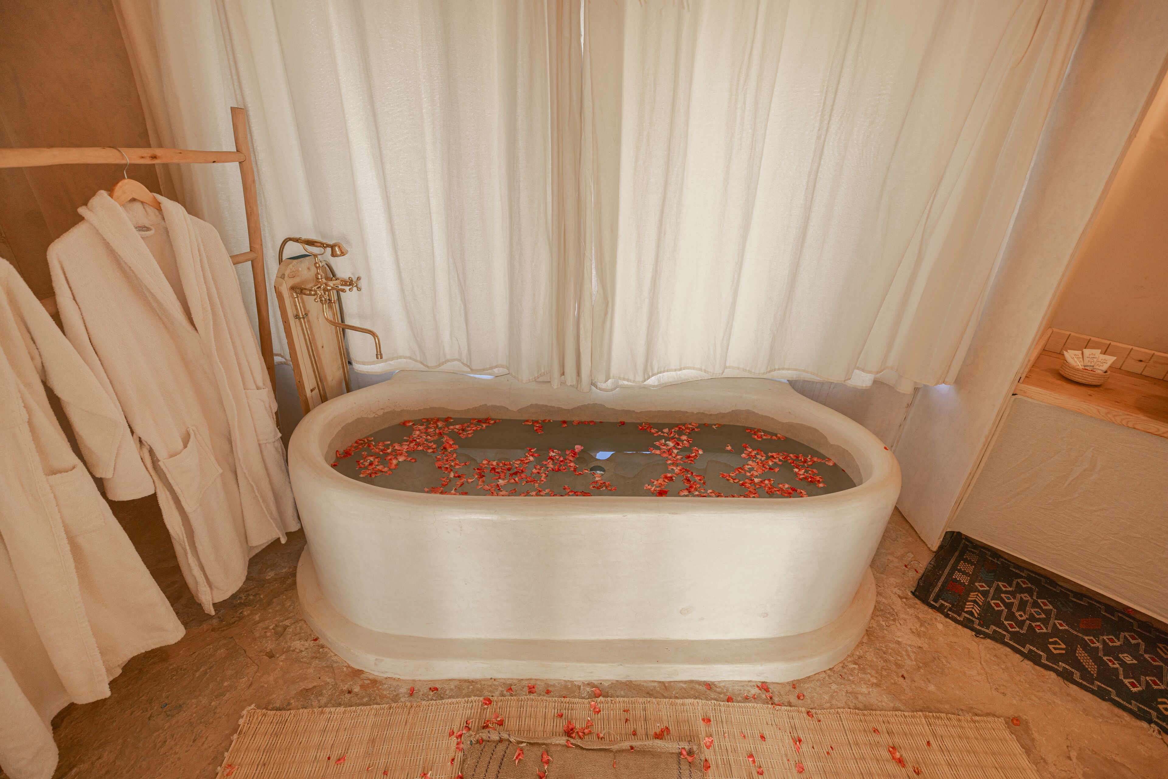 A bathtub filled with water and rose petals, surrounded by white towels and a wooden rack.
