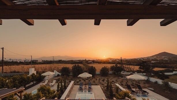 Área de piscina com espreguiçadeiras e guarda-sóis, com vista para um pôr do sol e uma montanha ao fundo.