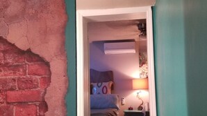 Un pasillo estrecho con una pared de ladrillo a un lado y una pared verde azulado al otro, que conduce a un dormitorio con cama, mesita de noche y lámpara.