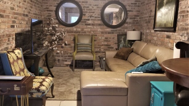 Una sala de estar acogedora con pared de ladrillo, dos espejos redondos, un sofá color beige, un sillón estampado, una mesita y un aparador verde azulado.