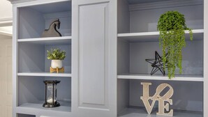 Uma estante branca com objetos decorativos, incluindo um vaso com plantas verdes, uma escultura geométrica preta e uma placa com a palavra 'LOVE'.