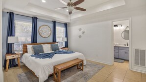 Quarto com cama, mesas de cabeceira, ventilador de teto e banheiro visível através de uma porta aberta.