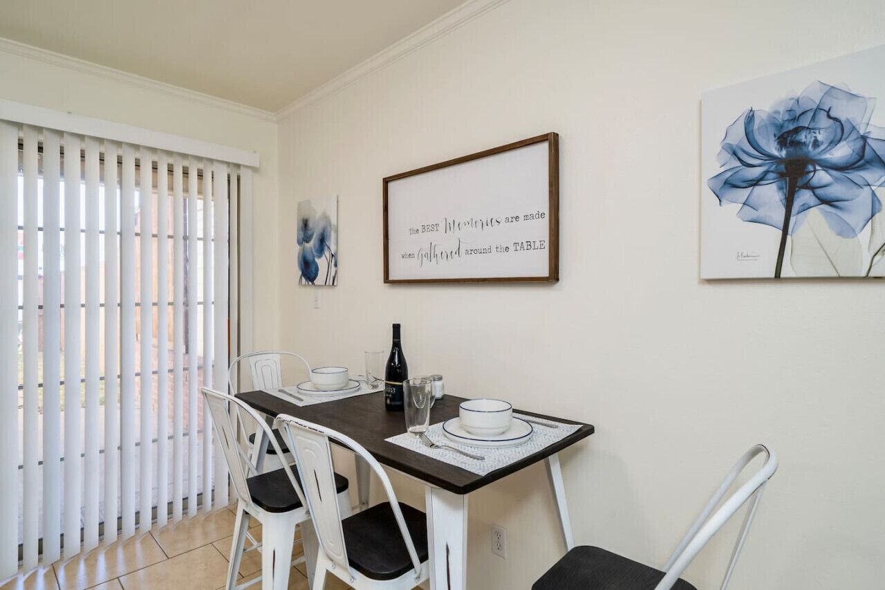 A dining area with a table set for two, white chairs, and framed artwork on the wall.