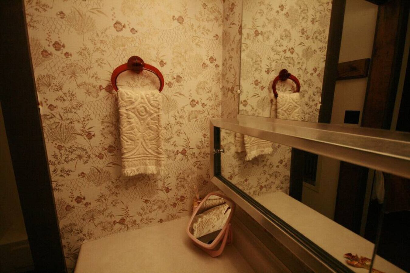 Ett badrum med blommig tapet, en handdukshängare med en handduk på och en spegel som reflekterar handduken.