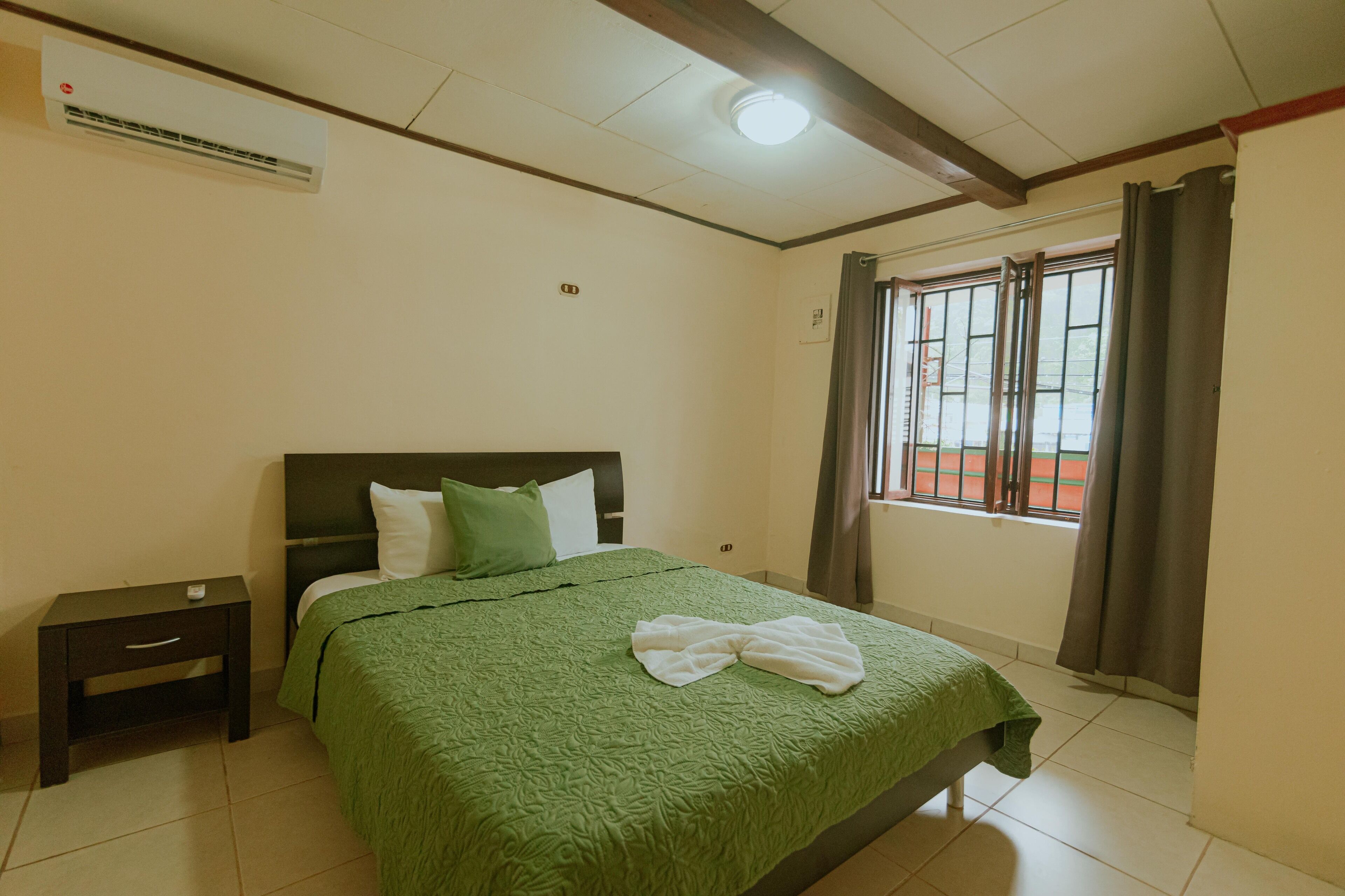 Un dormitorio con una cama, una mesita de noche, una ventana con cortinas y un equipo de aire acondicionado en la pared.