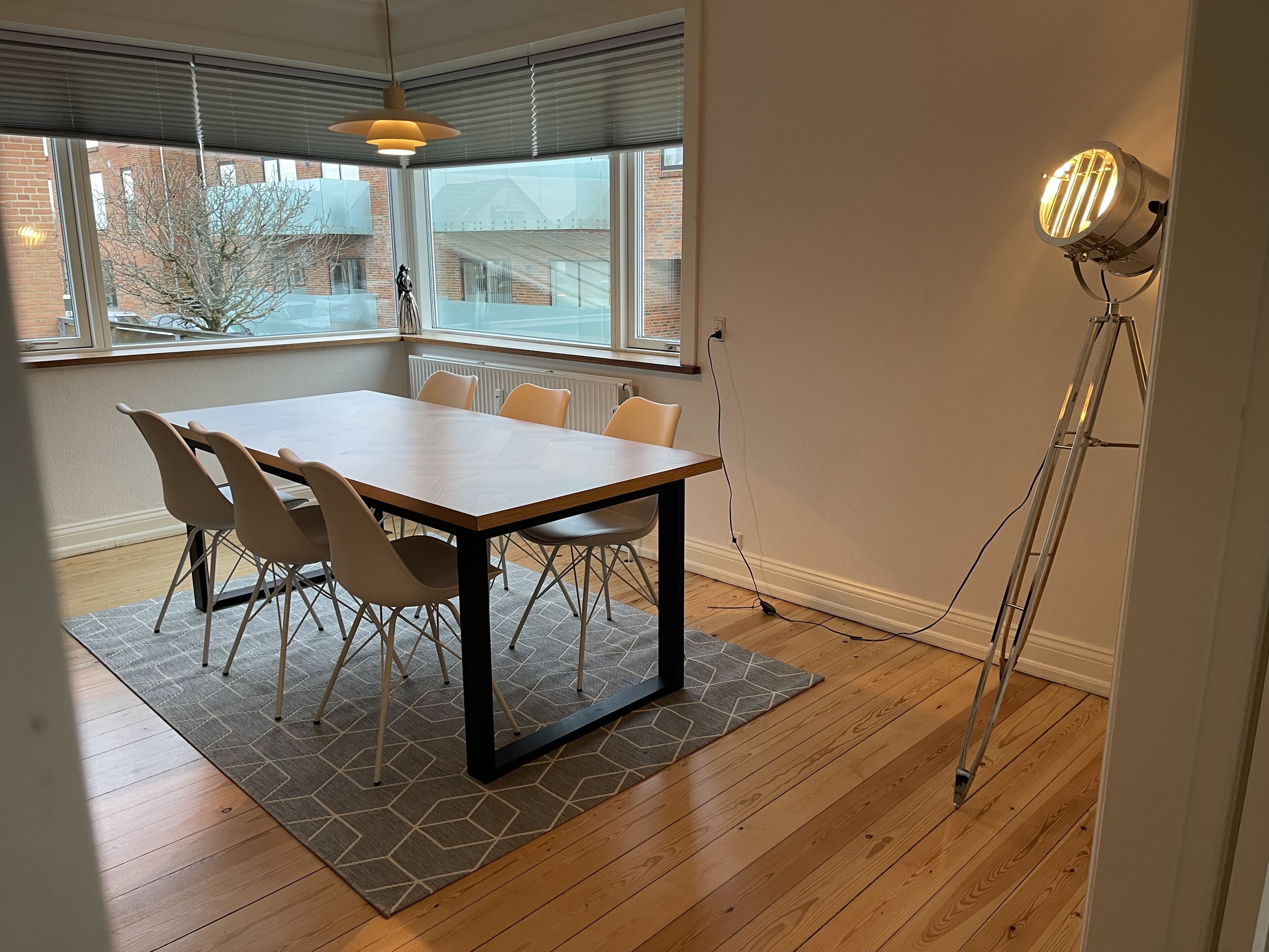 A conference room with a wooden table, six chairs, a patterned rug, and a large window with blinds.