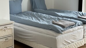 A bedroom with two beds, each with blue bedding and pillows. There is a bedside table with a lamp and a small dresser with baskets.