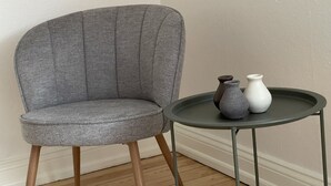 A grey upholstered armchair with wooden legs, positioned next to a small round table with two vases.
