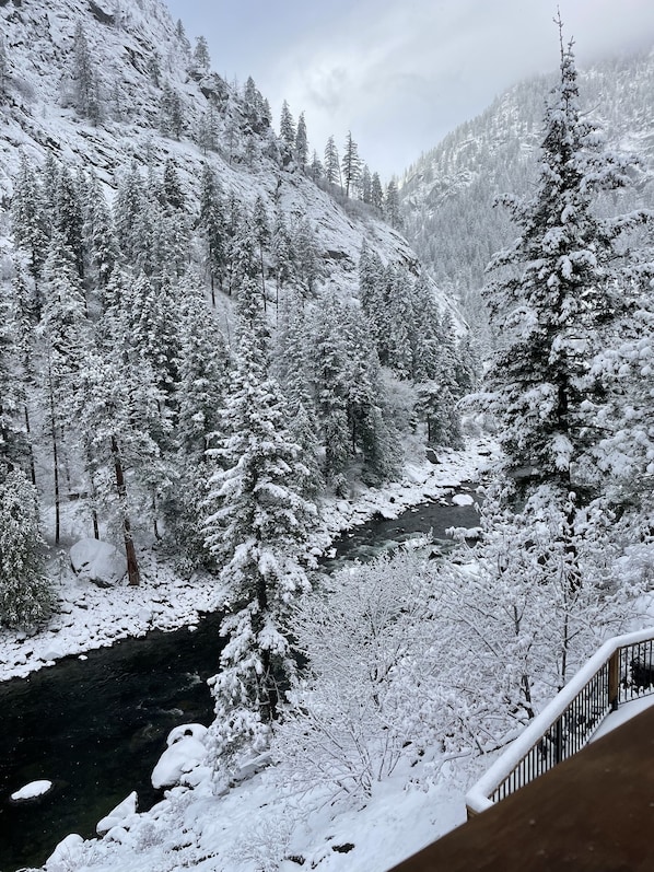 Paisaje montañoso nevado con un río, árboles y una terraza de madera.