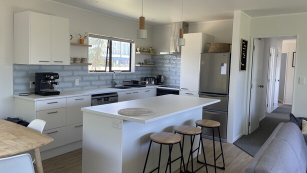 A modern kitchen with white cabinetry, a central island with stools, a coffee machine, and a refrigerator. - Ōhope Holiday Home  (Ōhope)