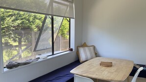 A room with a wooden table, white chairs, a bench, and a window with a view of greenery. - Ōhope Holiday Home  (Ōhope)