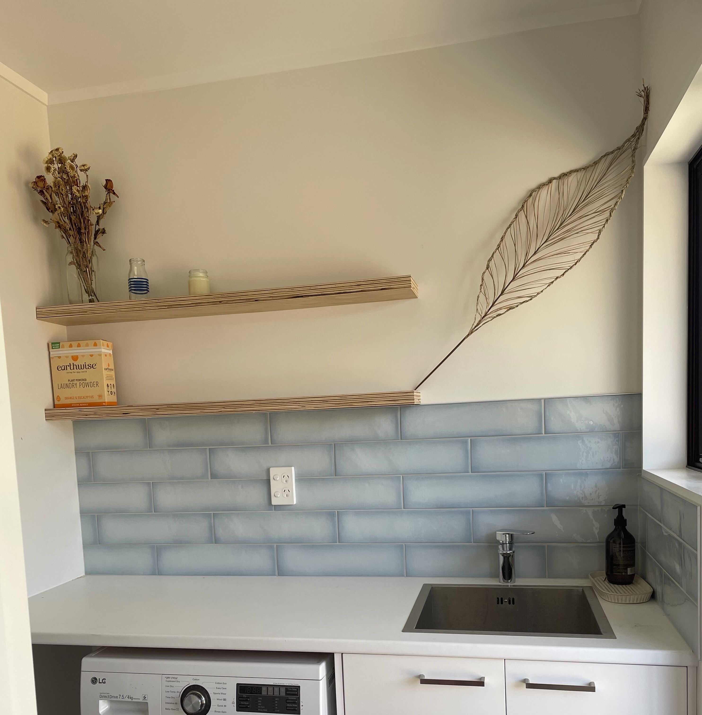 Een keuken met een wit aanrecht, een roestvrijstalen spoelbak en een wasmachine. Boven het aanrecht hangen twee planken; één met gedroogde bloemen en een fles, en de andere met een doos wasmiddel.