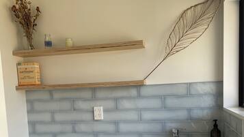 Een keuken met een wit aanrecht, een roestvrijstalen spoelbak en een wasmachine. Boven het aanrecht hangen twee planken; één met gedroogde bloemen en een fles, en de andere met een doos wasmiddel.