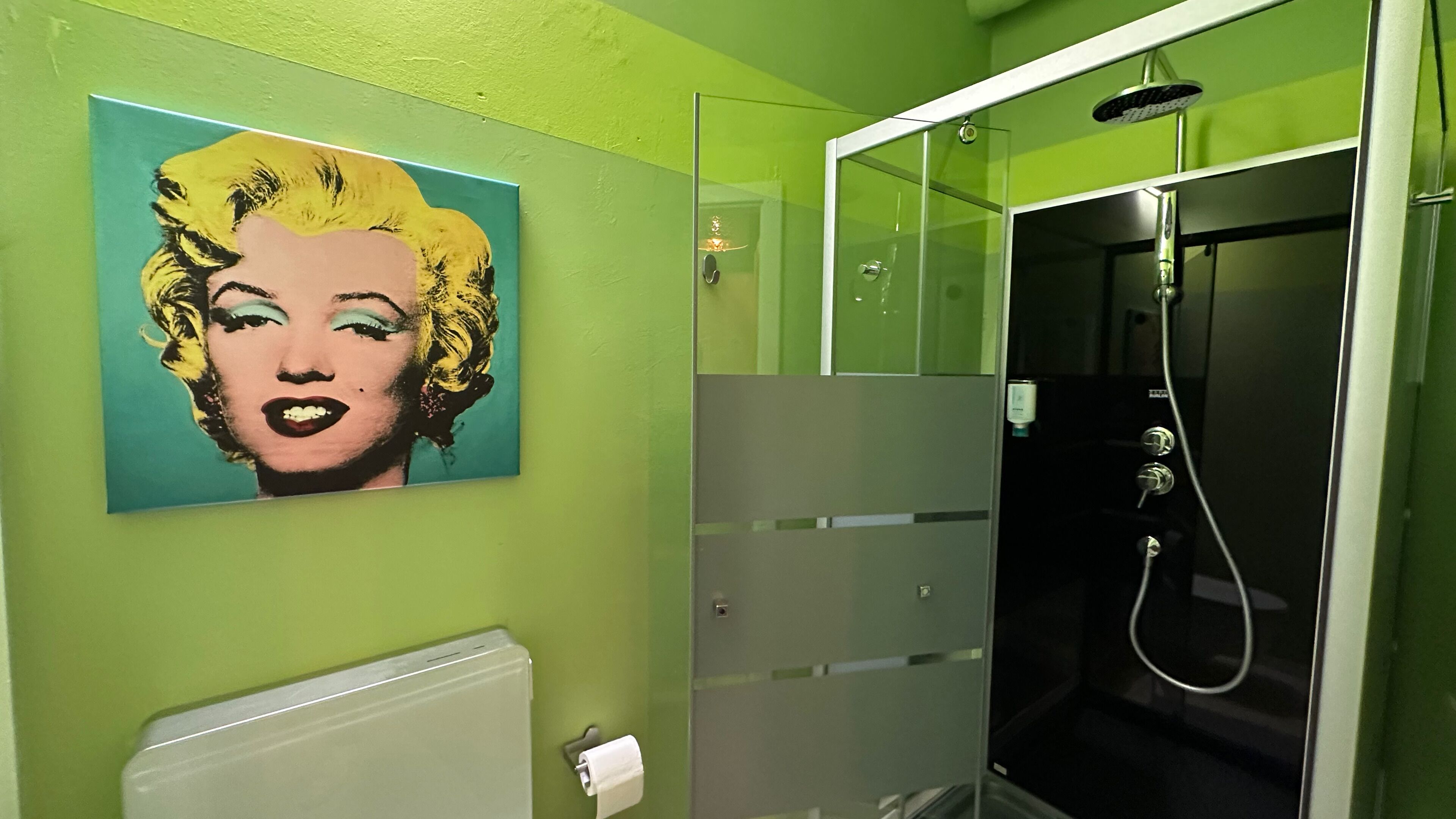 Un baño con pared verde, un cuadro de arte pop, cerramiento de ducha de vidrio y un radiador blanco.