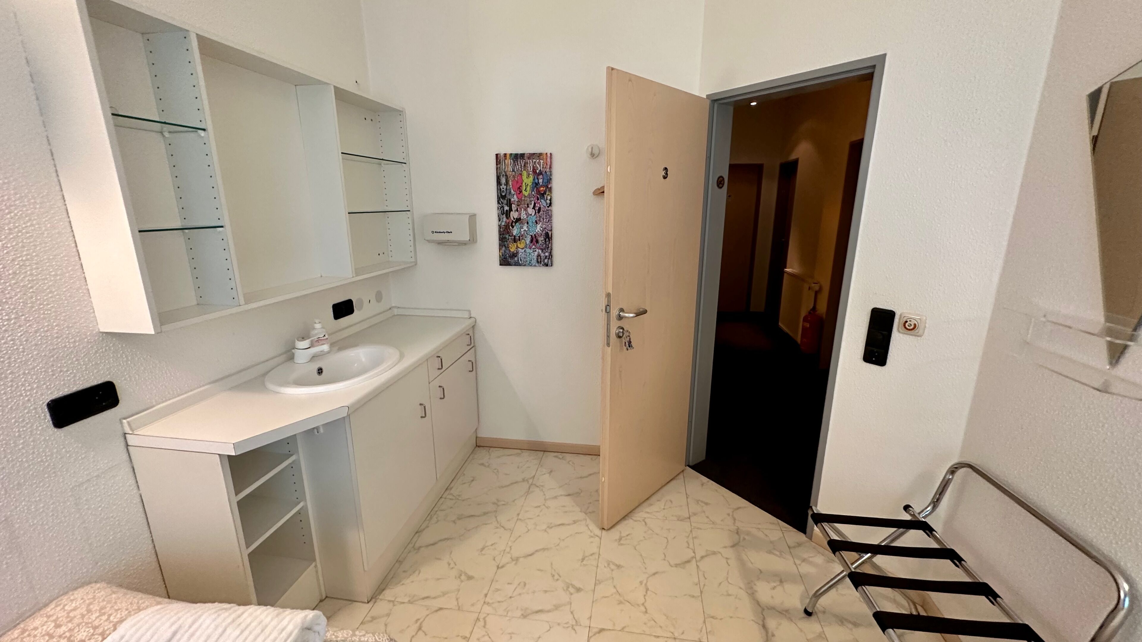 Een kleine, schone badkamer met een wit wastafel, spiegel en opbergkasten. Een deur leidt naar een gang met een brandblusser.
