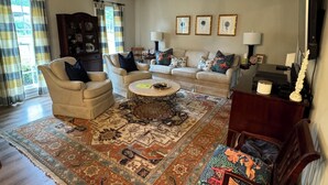 A living room with a beige sofa, armchairs, a coffee table, and a patterned rug. There are three framed pictures on the wall, a lamp, and a wooden cabinet.
