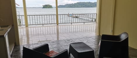 A balcony with a view of a lake and an island. There are two black chairs and a small table on the balcony floor.