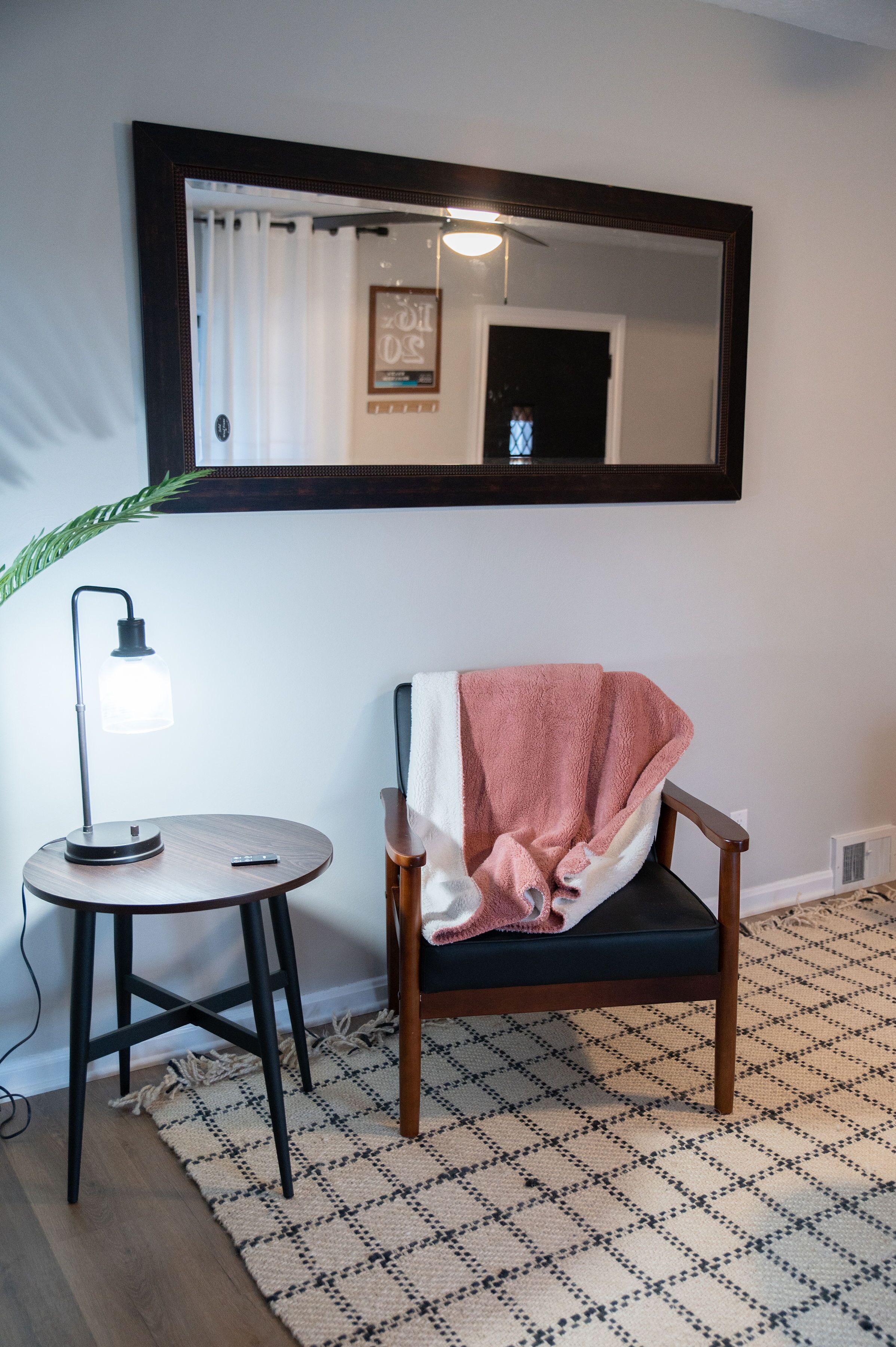 A room with a chair, a small table with a lamp, a patterned rug, and a large mirror on the wall.