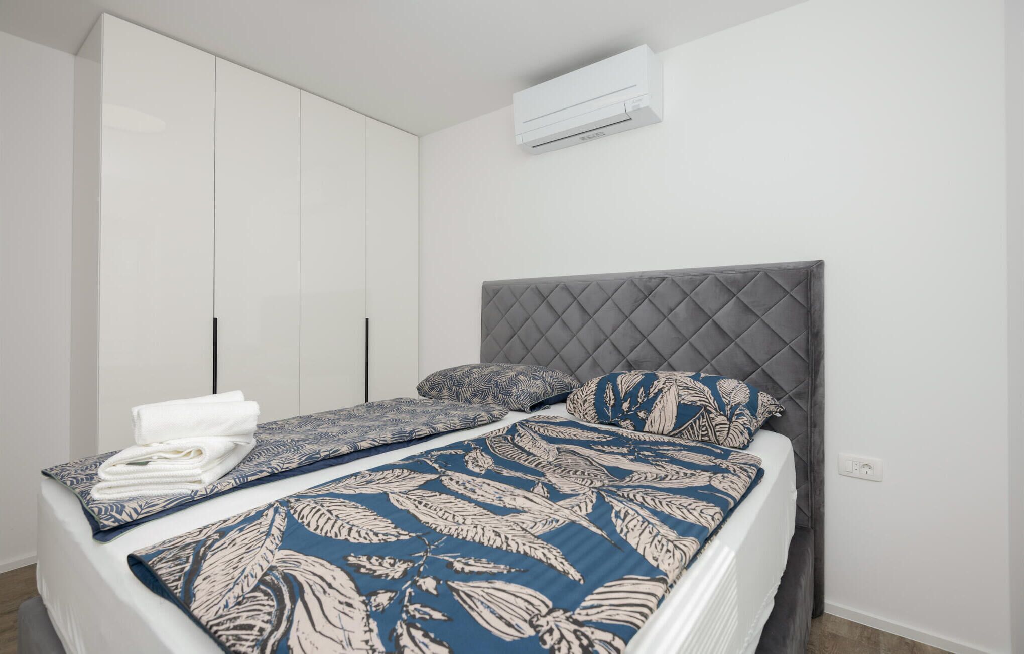 Un lit avec une couvre-lit à motifs, une tête de lit grise et un appareil de climatisation blanc placé au-dessus.