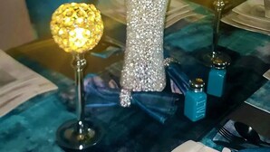 Un tavolo da pranzo con una tovaglia blu, un vaso decorativo con fiori artificiali, due lampade da tavolo e un paio di conditori.