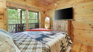 Ein Zimmer in einer Holzhütte mit einem Bett, einem Fernseher und einem Fenster mit Blick auf Grünfläche.