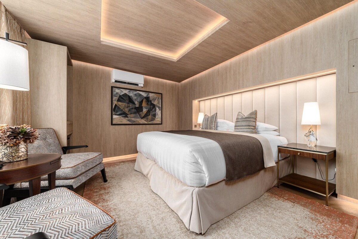 A modern hotel room with a large bed, a round table, a chair, and a bedside table with a lamp.