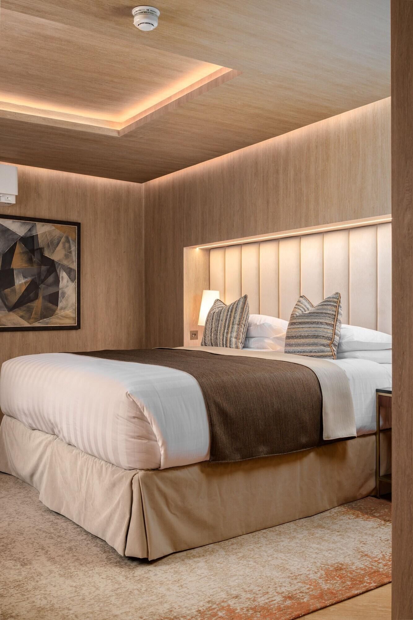 A modern bedroom with a large bed, a headboard with vertical panels, a wall-mounted light fixture, and a geometric artwork.