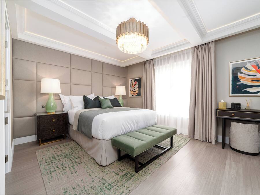 A modern bedroom with a large bed, bedside lamps, a bench, a desk, and a painting.