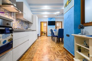 Un couloir étroit, avec une cuisine d’un côté, un coin repas avec des chaises bleues, et un téléviseur fixé au mur.