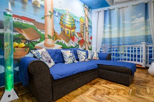 A cozy room with a blue sofa, a mural of a coastal scene, and a view of the sea through a window.