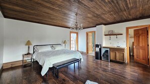A spacious bedroom with a large bed, a bench, a desk, and a kitchenette. - Nature-Filled Family Retreat Near Ann Arbor I Gardens, Bonfire & Wildlife (Ann Arbor)