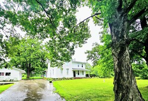 A white house with a front porch, surrounded by lush green trees and a well-maintained lawn. - Nature-Filled Family Retreat Near Ann Arbor I Gardens, Bonfire & Wildlife (Ann Arbor)