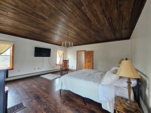A bedroom with a wooden ceiling, a bed with white bedding, a chandelier, a wooden chair, a television mounted on the wall, and a door. - Nature-Filled Family Retreat Near Ann Arbor I Gardens, Bonfire & Wildlife (Ann Arbor)