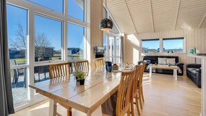 Een ruime woonkamer met een houten eettafel, stoelen en een bank. Grote ramen bieden uitzicht op het omliggende landschap.