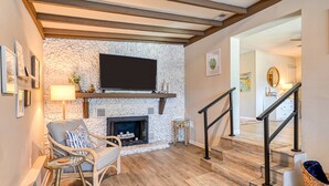 A living room with a stone fireplace, a flat-screen TV mounted above it, a wicker chair, and a staircase leading to another room. - The Pelican Point (St. Augustine)