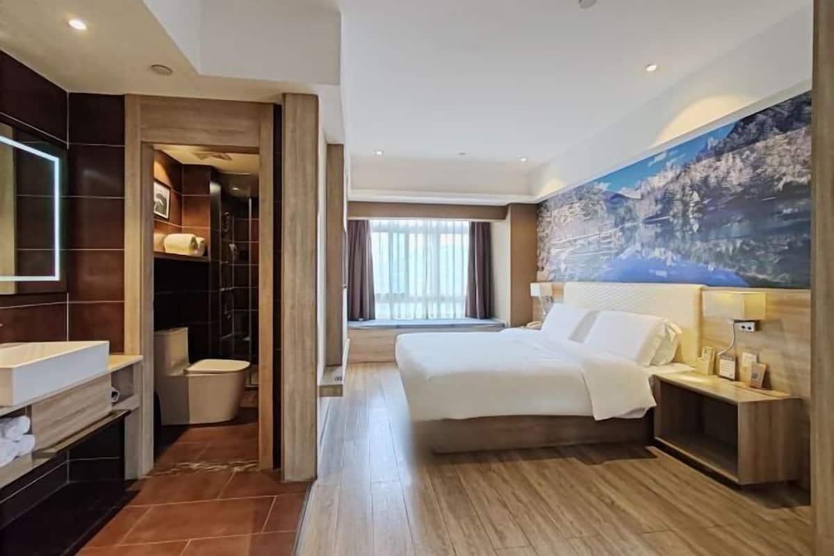 A hotel room with a large bed, a bathroom with a shower, a vanity, and a large window.