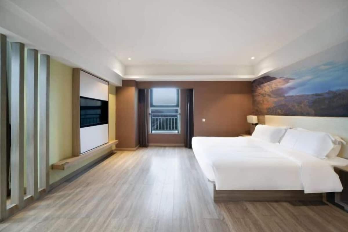 A hotel room with a bed, a TV, a window, and a painting on the wall.