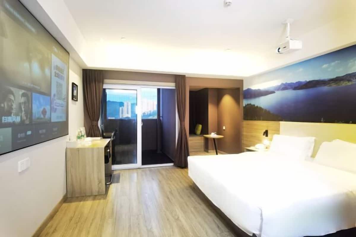A hotel room with a bed, a desk, a chair, a small table, a projector, and a large window.