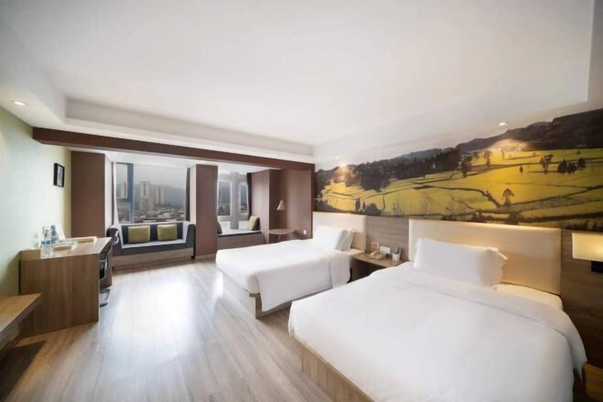 A hotel room with two beds, a desk, a window with a city view, and a large landscape painting on the wall.